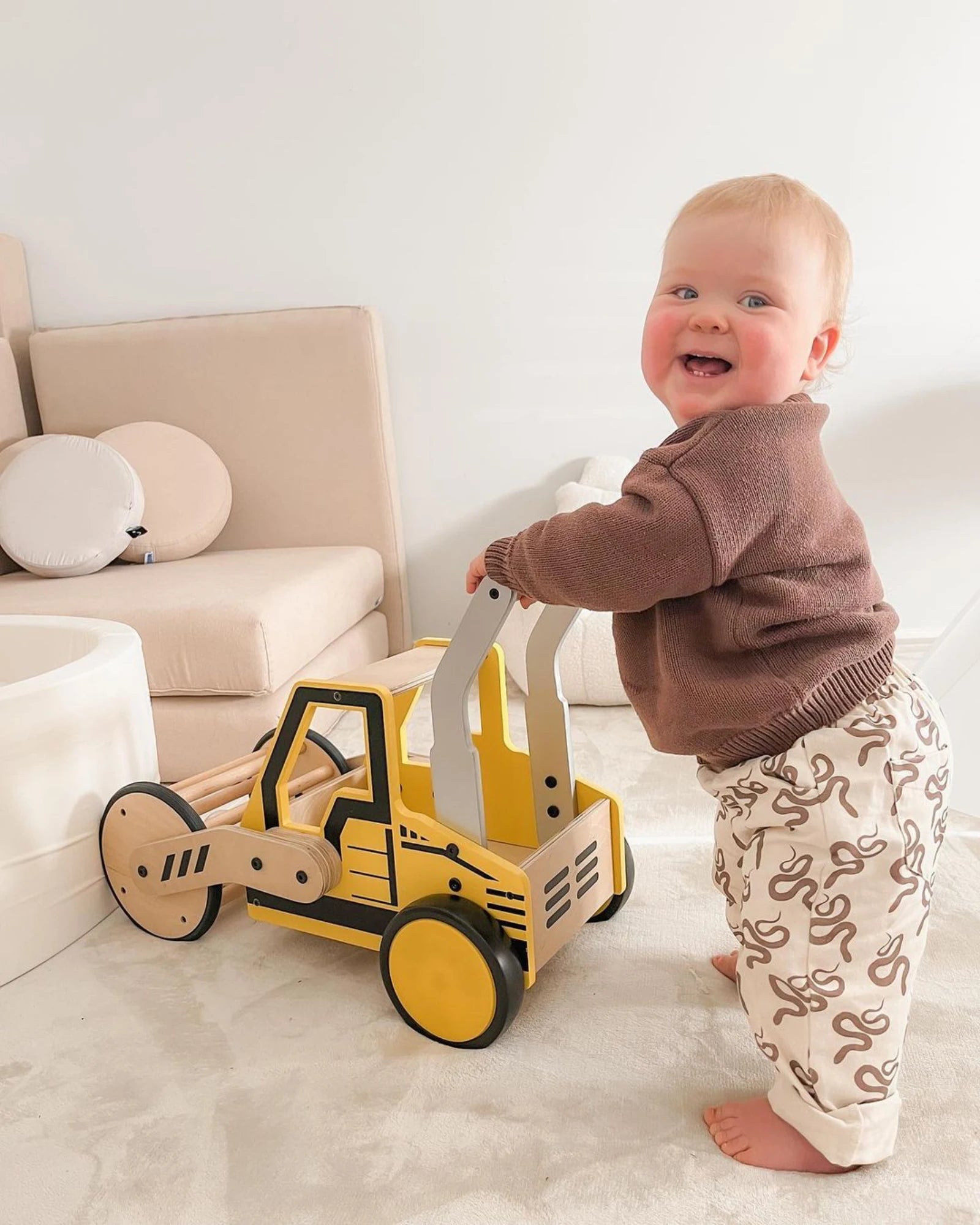 A smiling toddler holding the Riababy Road Roller push walker in a bright family room
